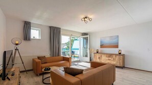 A modern living room with a sofa, a coffee table, a lamp, and a view of the outdoors. - Apartment by Dutch Beach Pet-friendly (Julianadorp)