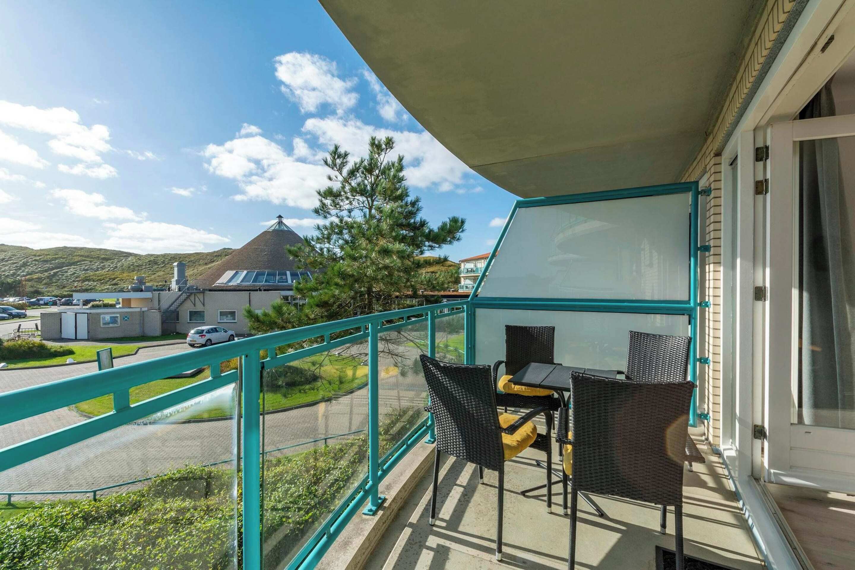 A balcony with two wicker chairs and a table, overlooking a parking area and a building with a conical roof.