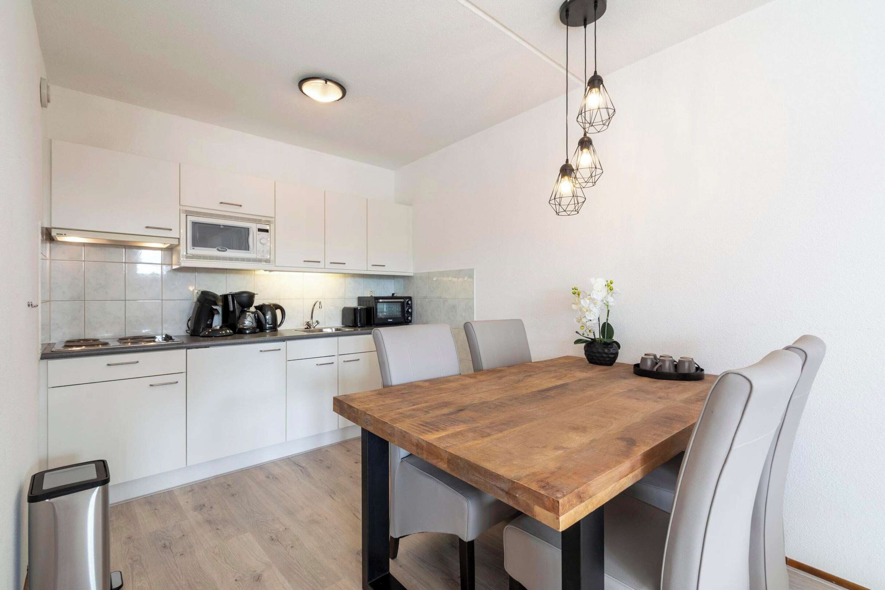 A modern kitchen with white cabinets, a wooden dining table, and a stainless steel trash bin.