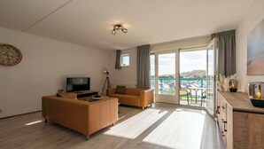 A modern living room with a sofa, TV, and a balcony with a view. - Apartment by Dutch Beach Pet-friendly (Julianadorp)
