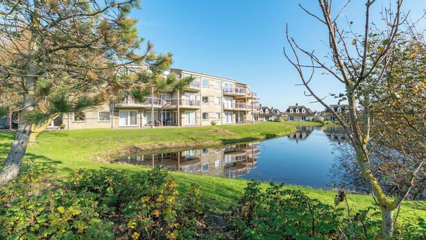 Un edificio residenziale a più piani con balconi, un piccolo stagno in primo piano e vegetazione circostante.