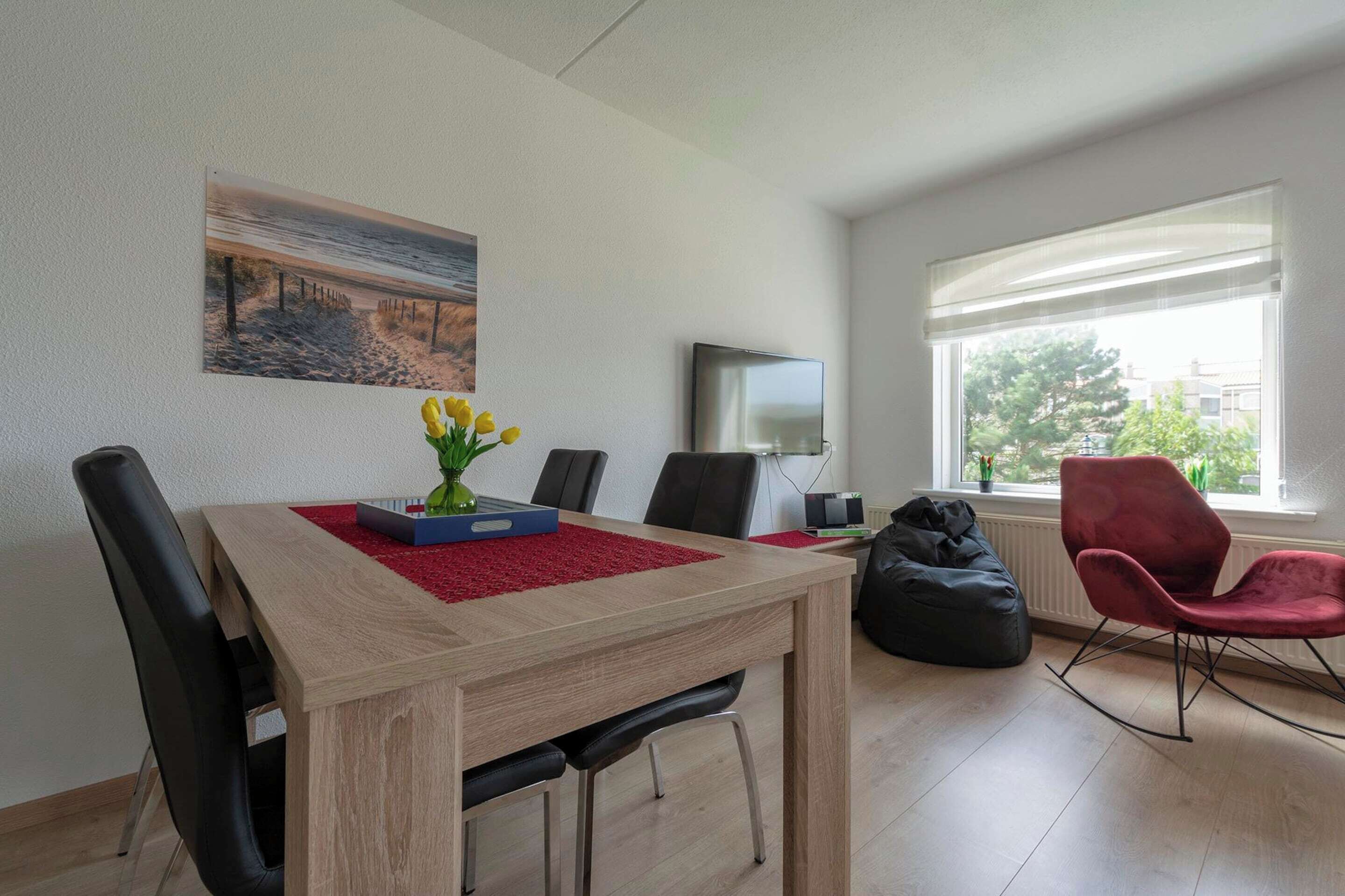 En moderne stue med et træspisebord, sorte stole, en rød velour-lænestol, en sort poseleje, et fladskærms-tv og et maleri af et strandmotiv.