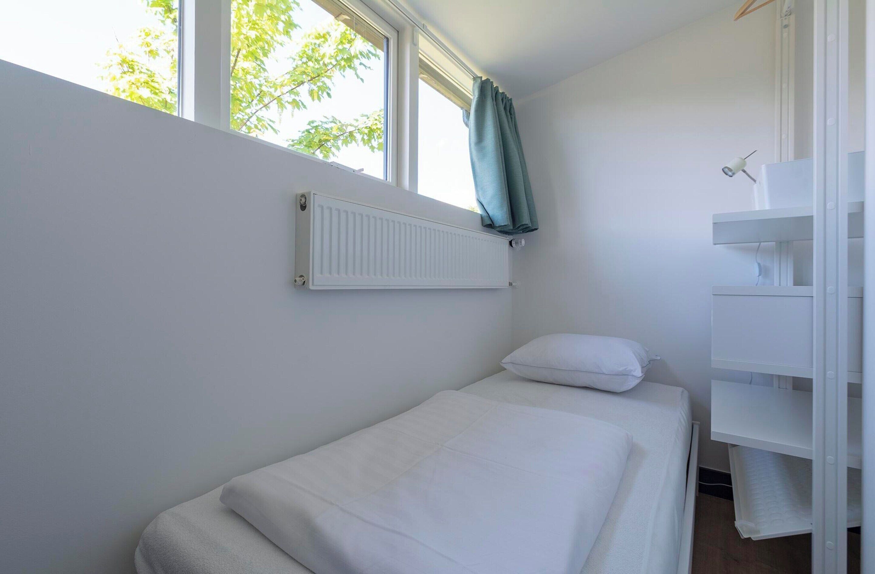 Une petite chambre avec un lit simple, une fenêtre, un radiateur et une étagère blanche.