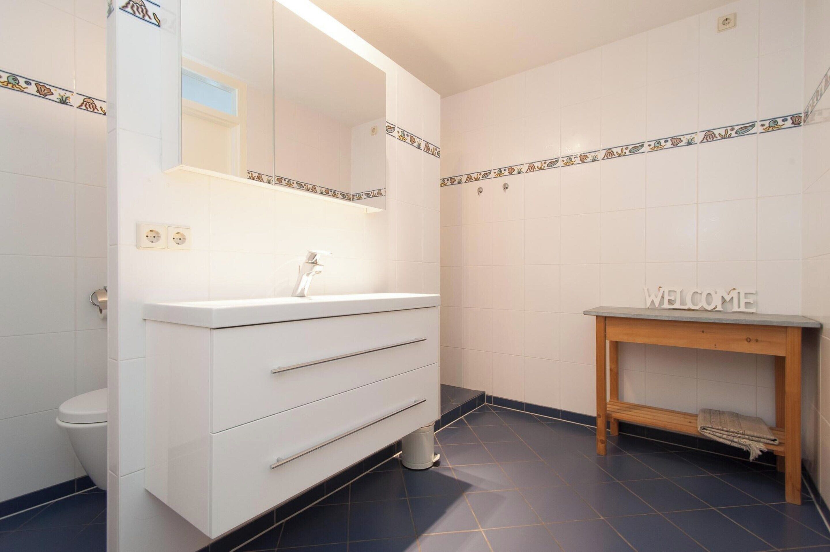 A bathroom with a white vanity, a mirror, a sink, a toilet, and a welcome sign on a wooden shelf.