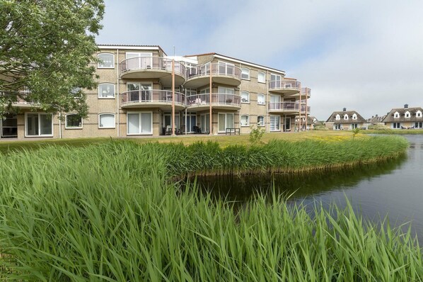 A multi-story residential building with balconies, a canal in front, and a grassy area. - Stunning Apartment Near sea (Julianadorp)
