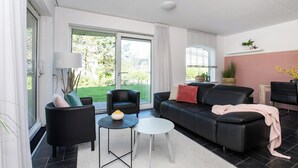 En moderne stue med en sort lædersofa, et hvidt sofabord og en sort lænestol. Et stort vindue med udsigt over en grøn have.