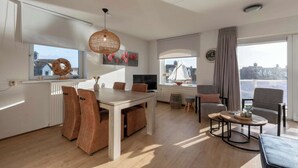 Ein modernes Wohnzimmer mit Essbereich, großen Fenstern mit Vorhängen, einem Flachbildfernseher und Blick auf die Stadt.