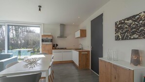 A modern kitchen with white cabinetry, a wooden countertop, and a built-in stove. There is a dining area with a white table and chairs, and a large window providing a view of the outdoors. - Modern Coastal Escape (Sint Maartensvlotbrug)