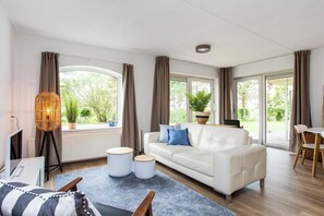 Un soggiorno moderno con un divano bianco, pouf rotondi, un'ampia finestra con tende, una lampada da terra e vista su vegetazione.