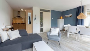 A modern living room with a grey sofa, a small armchair, and a dining table with white chairs. There is a kitchen area visible in the background. - Apartment in Julianadorp Near the Beach (Julianadorp)