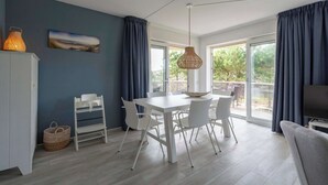 A modern kitchen with white cabinetry, a dining table with chairs, a hanging light fixture, and a large window with curtains. - Apartment in Julianadorp Near the Beach (Julianadorp)