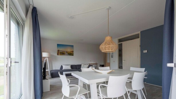 A modern dining area with a white table and chairs, a hanging wicker light fixture, and a view of the outdoors through large windows. - Apartment in Julianadorp Near the Beach (Julianadorp)