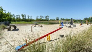 A playground with a slide, swings, and a seesaw on a sandy area with a picnic table and benches nearby. - Seaside Getaway With Terrace (Callantsoog)