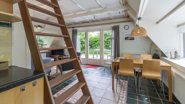 A kitchen with a wooden ladder, a dining area with chairs and a table, and a view of the outdoors through large windows. - Seaside Getaway With Terrace (Callantsoog)