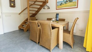 Een eetgedeelte met een houten tafel, rieten stoelen, een spiltrap, een klok en een schilderij van een vuurtoren.