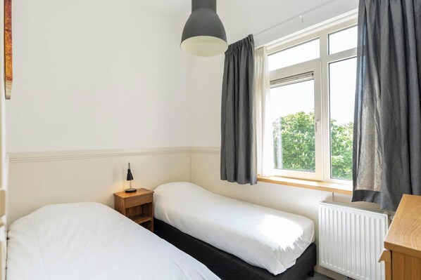 A bedroom with two beds, a bedside table with a lamp, a window with curtains, and a radiator. - Holiday Home in De Koog Near Beach (De Koog)