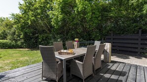 Een buitenterras met een houten dek, rieten stoelen en een tafel met een mand en flessen.