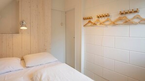 A bedroom with a wooden headboard, a bed with white linens, a wall-mounted light fixture, and a wooden coat rack with hangers. - Holiday Home Groote Keeten Near Beach (Callantsoog)