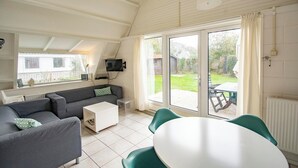 A modern living room with a sofa, coffee table, and a dining table with chairs. There is a television mounted on the wall, and a sliding glass door leading to an outdoor patio. - Holiday Home Groote Keeten Near Beach (Callantsoog)