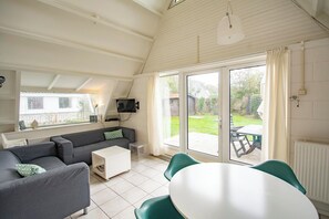 A modern living room with a sofa, coffee table, and a dining table with chairs. There is a television mounted on the wall, and a sliding glass door leading to an outdoor patio.