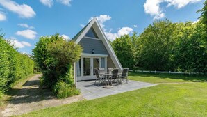 Uma casa pequena e moderna com telhado triangular, um pátio com mesa e cadeiras e um gramado bem cuidado.