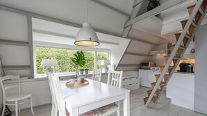 Cozinha moderna com armários brancos, mesa de jantar com cadeiras, uma janela grande com vista para o verde e uma escada de madeira.