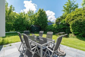 A patio with a glass-top table and six chairs, surrounded by lush greenery and a small shed. - Sandepark 165 Holiday Home Near Beach (Callantsoog)