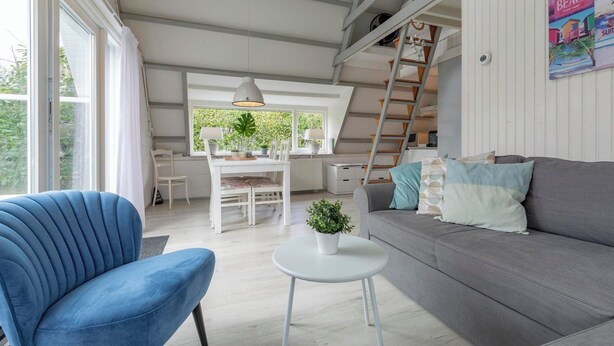 Uma sala de estar moderna com uma cadeira azul, um sofá cinzento e uma pequena mesa branca com uma planta. Há uma escadaria que leva a um andar superior, e uma secretária branca com uma cadeira e uma planta ao fundo.