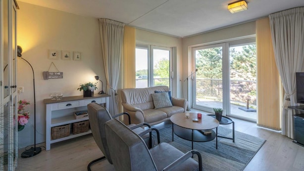 A modern living room with a sofa, chairs, a coffee table, and a TV. There are large windows with curtains, a small table with a plant, and a wall-mounted shelf with decorative items. - Cozy Dune-view Apartment (Julianadorp)