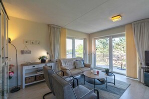 A modern living room with a sofa, chairs, a coffee table, and a TV. There are large windows with curtains, a small table with a plant, and a wall-mounted shelf with decorative items. - Cozy Dune-view Apartment (Julianadorp)