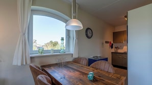 A dining area with a wooden table, chairs, a hanging light fixture, a window with curtains, and a view of the outside. - Cozy Dune-view Apartment (Julianadorp)