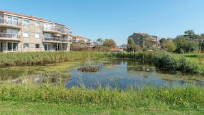 A residential area with modern apartment buildings, a pond with a small island, and surrounding greenery. - Cozy Dune-view Apartment (Julianadorp)