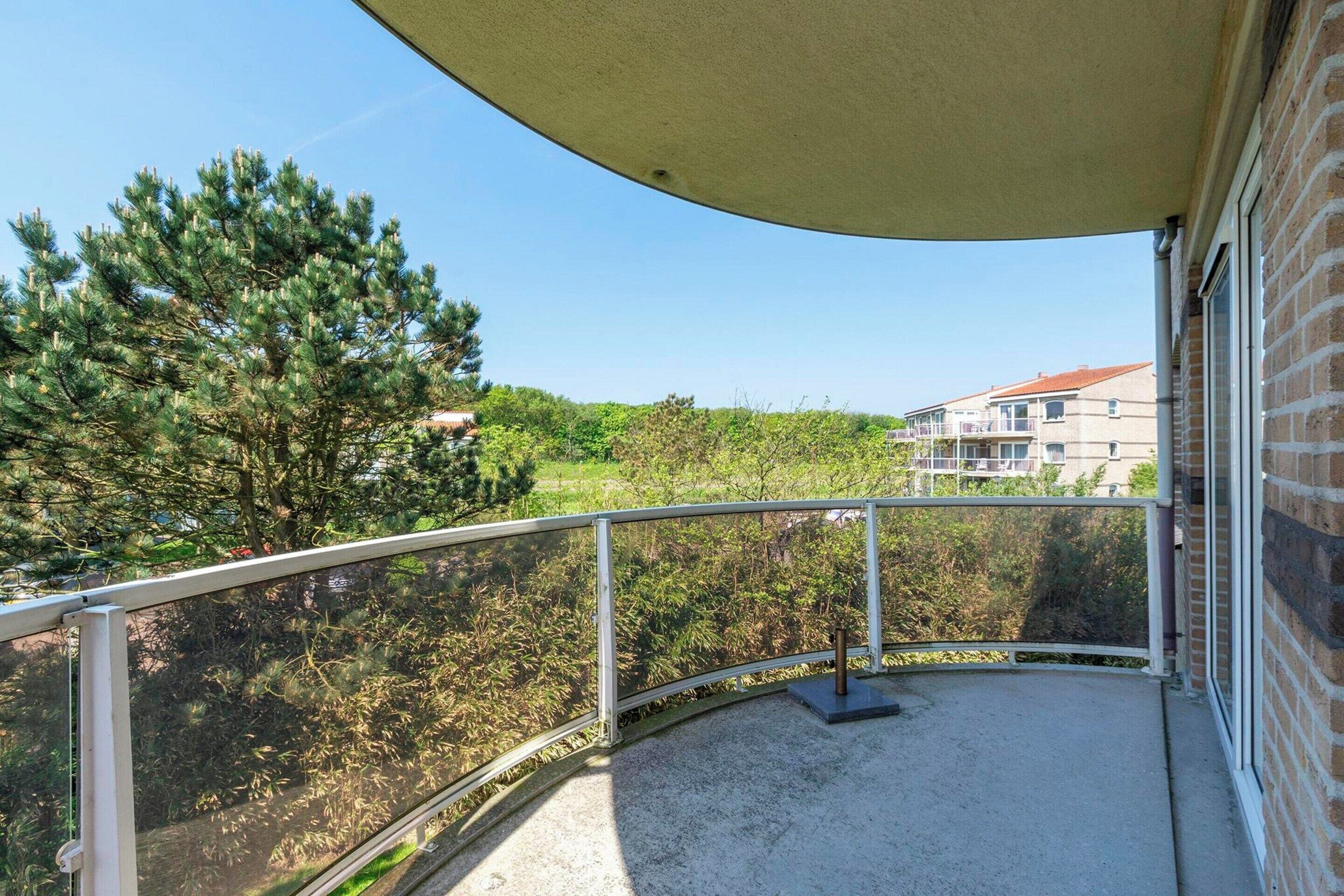 Un balcón con baranda metálica, piso de concreto y vista a árboles y edificios al fondo.
