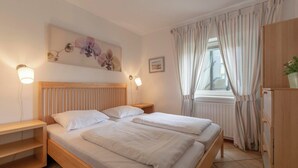 A double bed with a wooden headboard, two bedside tables, a small shelf, a wall-mounted lamp, a window with curtains, and a framed picture.