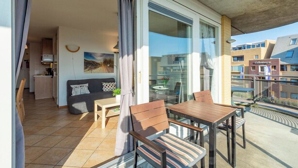 A balcony with a dining table and chairs, a sofa, and a kitchen area visible through the glass doors.