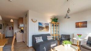 A modern living room with a grey sofa, a wooden dining table, and a small white table with a plant. There is a kitchen in the background with a white fridge and cabinets.