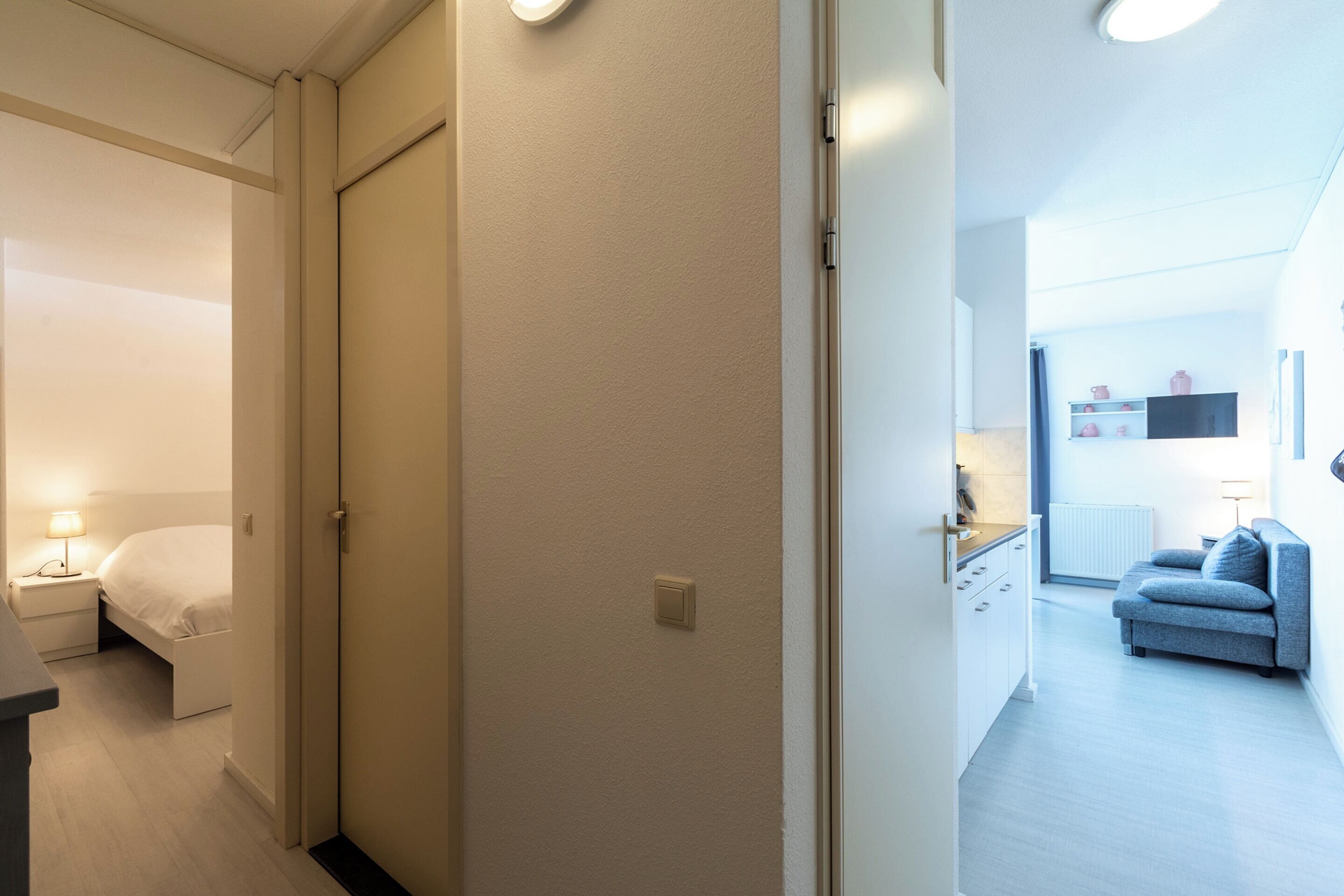 Et hotelværelse med en seng, et natbord og en lampe. Der er en dør, der fører til et andet rum.