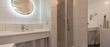 A modern bathroom with a white sink, a round mirror with lights, a glass-enclosed shower, and a toilet.