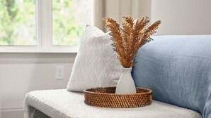 A room with a window, a white textured bench, a blue sofa, a woven basket, and a vase with dried plants.