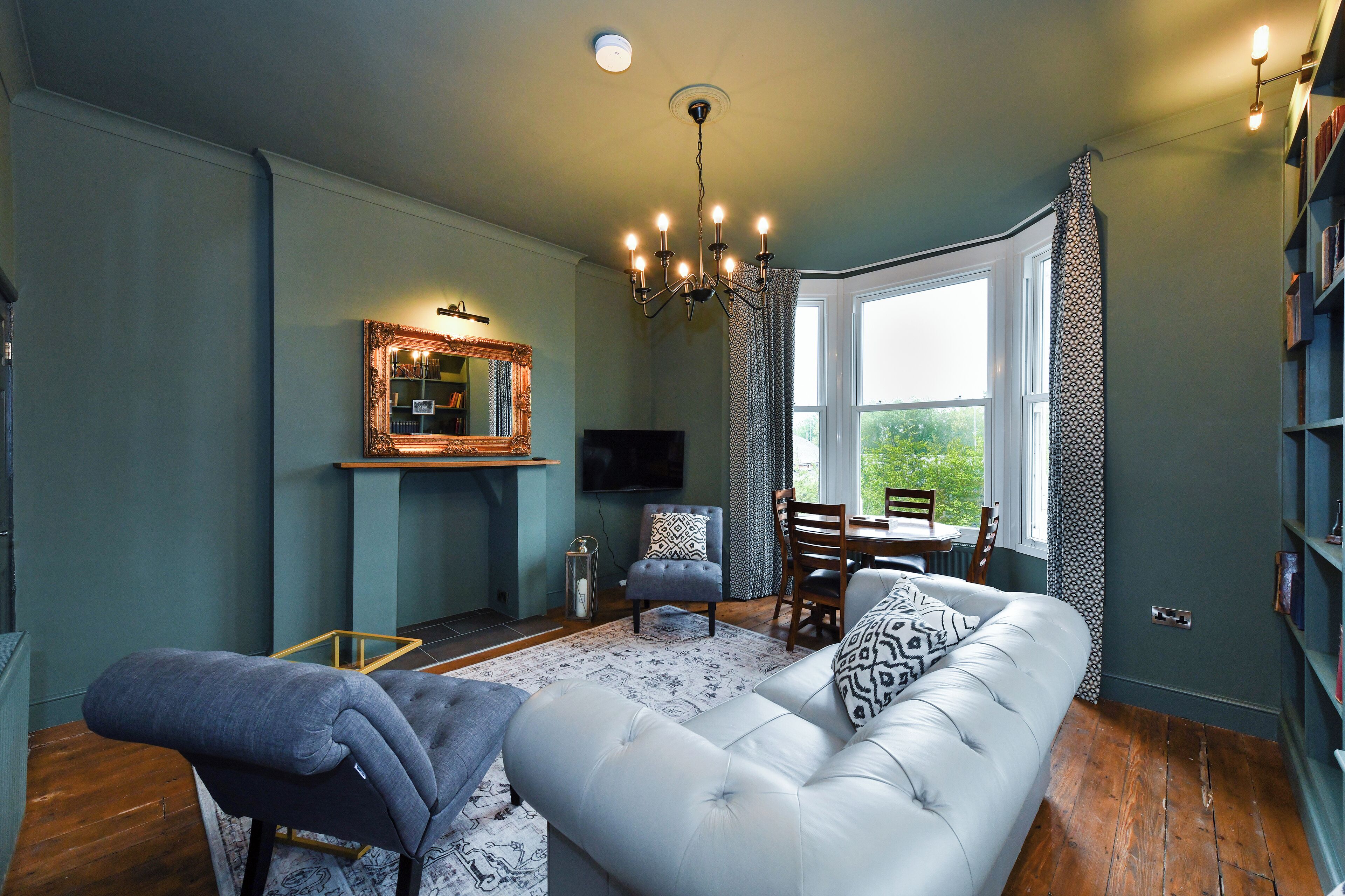 A living room with a large grey sofa, a patterned rug, a chandelier, a fireplace, and a bookshelf.