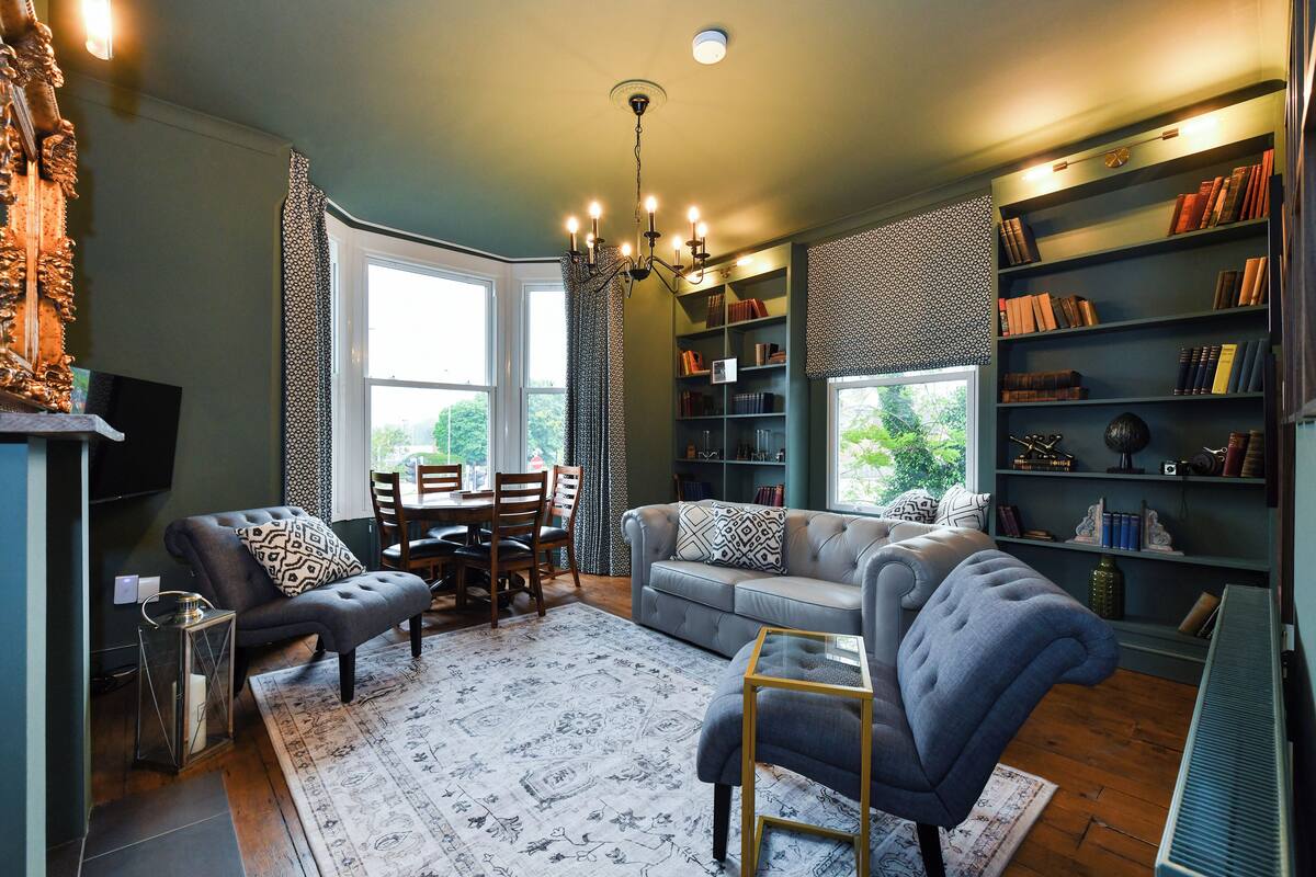 A living room with a sofa, armchair, and a dining table with chairs. There is a bookshelf filled with books and a chandelier.