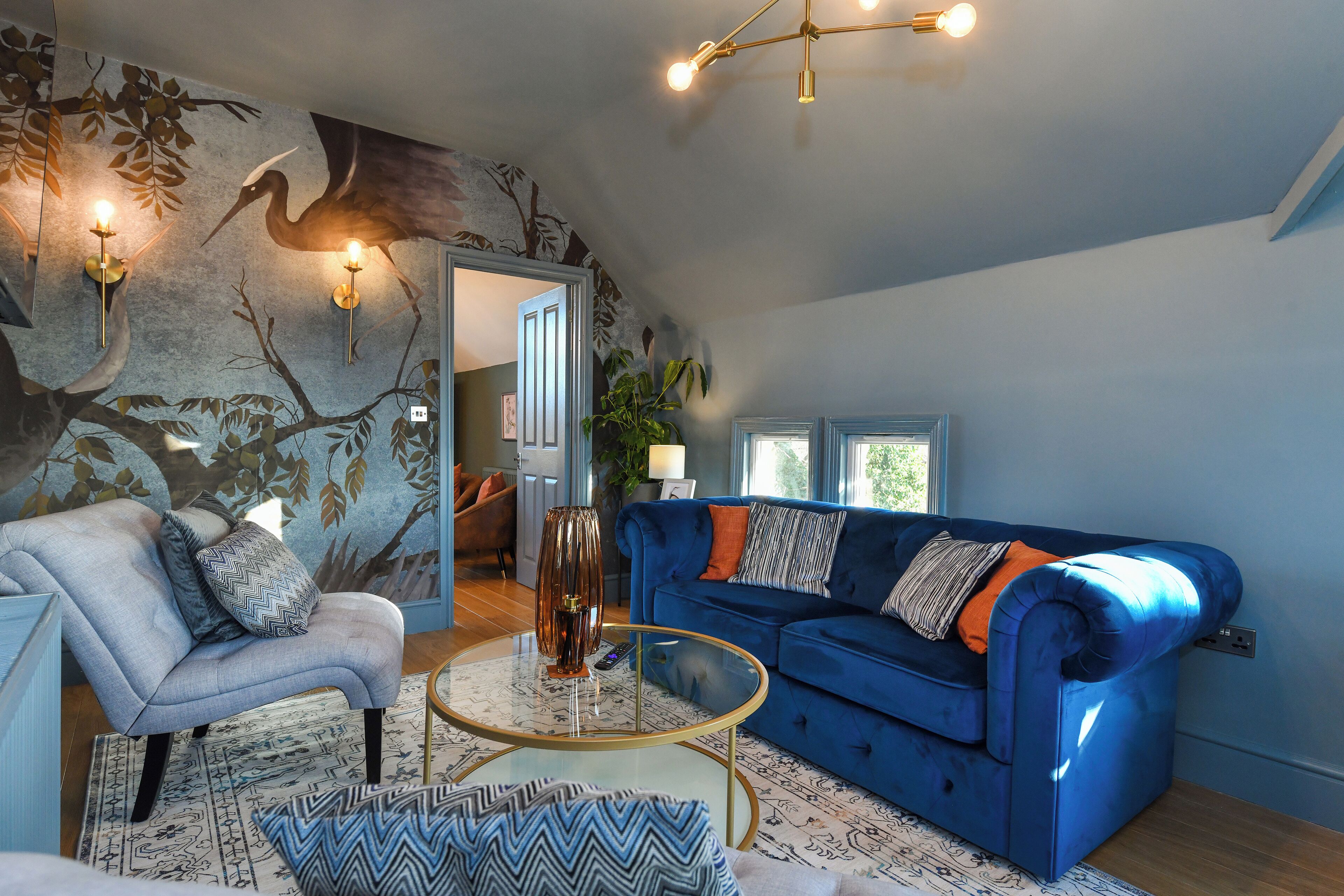 Un salon avec un canapé bleu, un fauteuil gris et une table basse en verre. On y trouve des appliques murales avec des lampes, une fresque sur le mur et une porte qui donne sur une autre pièce.