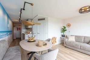 A modern living room with a round dining table, grey sofa, and a view of the sea through a window. - Stylish Couples' Retreat (Callantsoog)