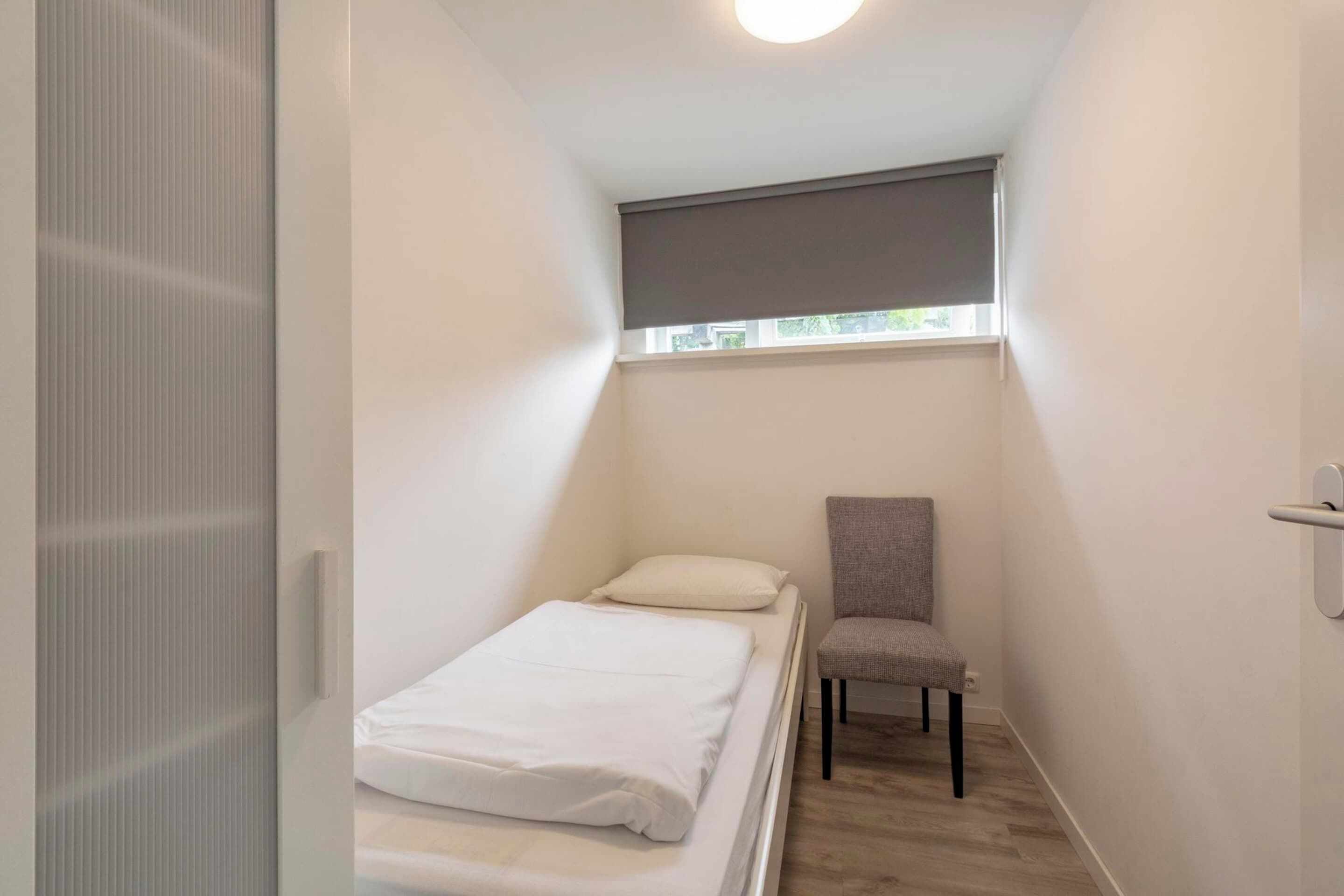 A small, narrow room with a single bed, a chair, and a window with a blind.