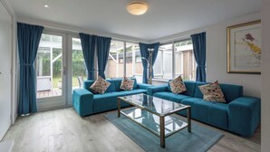 A spacious living room with a teal sofa set, a glass coffee table, and large windows with blue curtains. - Bungalow in Sint Maartenszee Near Beach (Sint Maartensvlotbrug)