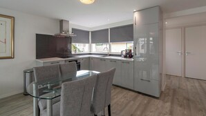 A modern kitchen with a glass dining table, grey upholstered chairs, and white cabinetry. - Bungalow in Sint Maartenszee Near Beach (Sint Maartensvlotbrug)