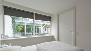 A bedroom with a bed, a window with a view of a house and greenery, and a bedside table with a lamp. - Bungalow in Sint Maartenszee Near Beach (Sint Maartensvlotbrug)