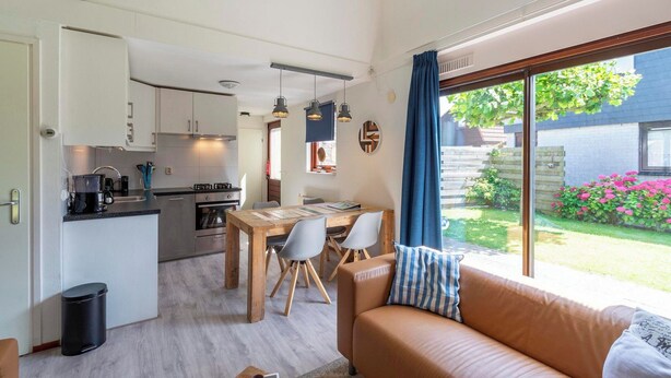 A compact living space with a kitchenette, dining area, and a large window offering a view of a garden. - Yperhof 66 Holiday Home for 6 Guests on Cost (Julianadorp)