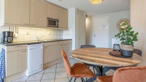 Une cuisine moderne avec des meubles en bois, un four à micro-ondes et un lave-vaisselle. On y trouve une table ronde en bois, des chaises et une plante en pot.