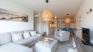 A modern living room with a sofa, ottoman, and coffee table, a dining area, and a kitchen in the background. - Wijde Blick 212 Apartment in Callanstoog (Callantsoog)
